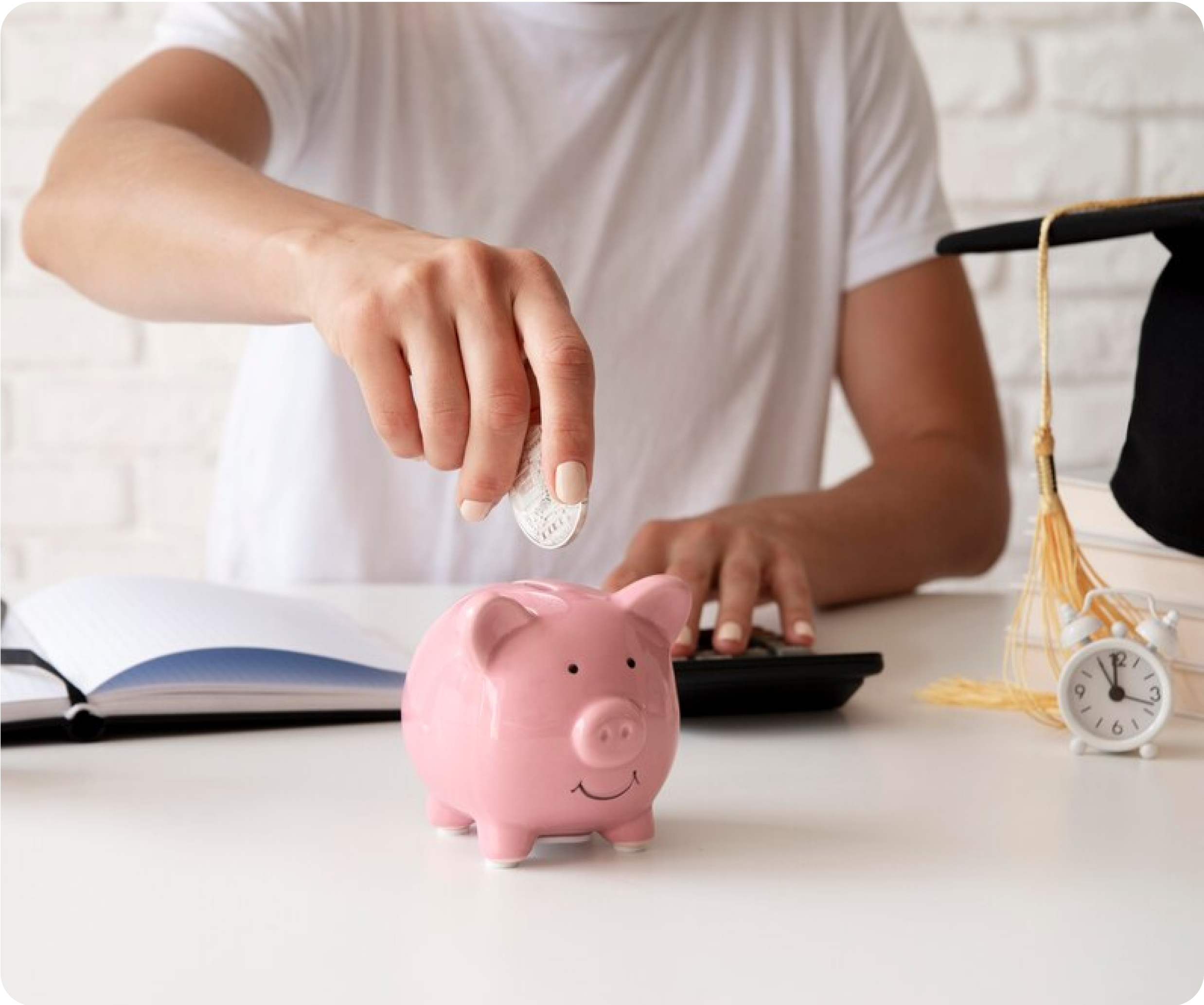 Person putting coin into a pink piggy bank with a graduation cap and book nearby.