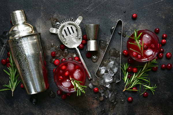 Cocktail shaker, strainer, jigger, and ice tongs on a black slate background, with a red, cranberry and rosemary garnished cocktail, and several cranberries, sprigs of rosemary, and pieces of crushed ice strewn around artfully 
