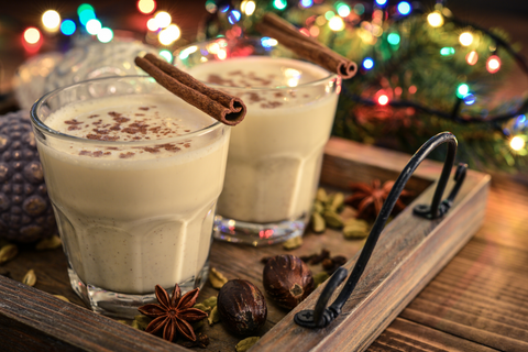 Two glasses of eggnog, garnished with grated nutmeg and cinnamon sticks, on a wooden tray, with multi-colored Christmas tree lights in the background