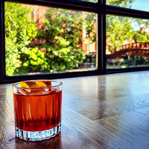 A Boulevardier cocktail on a wooden bar, with a window in the background showing a sunny day with trees and a bridge