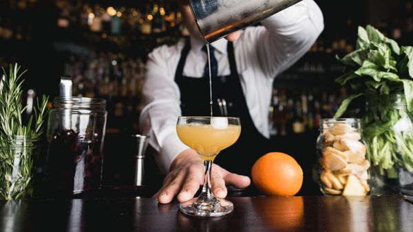 A bartender in a white shirt and black leather apron pours an orange cocktail into a cut crystal coupe glass