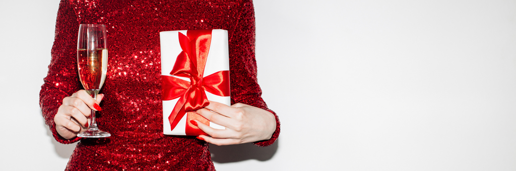 Picture of a woman's torso in a red sequined dress holding a champagne flute in one hand and a white gift with a red ribbon in the other, against a white background