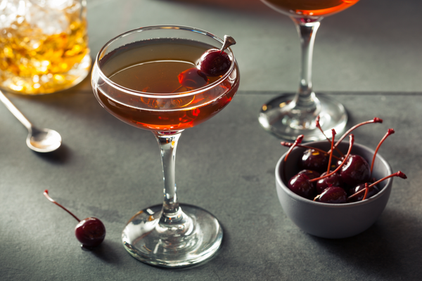 A crystal mixing glass, silver bar spoon, two Manhattan cocktails in coupe glasses, and a small ramekin of cherries on a gray slate surface.