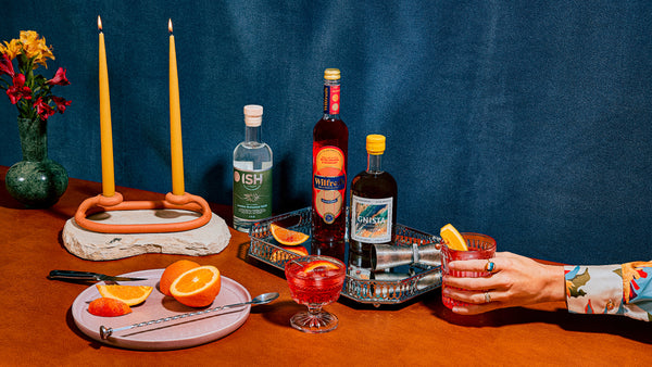 A table set with an orange table cloth, two yellow taper candles, a bouquet of flowers, a silver tray, a plate of sliced oranges, a Negroni cocktail, bottles of GinISH, Wilfred's Bittersweet Aperitif, and Gnista Floral Wormwood. A hand reaches from the right side holding another Negroni.