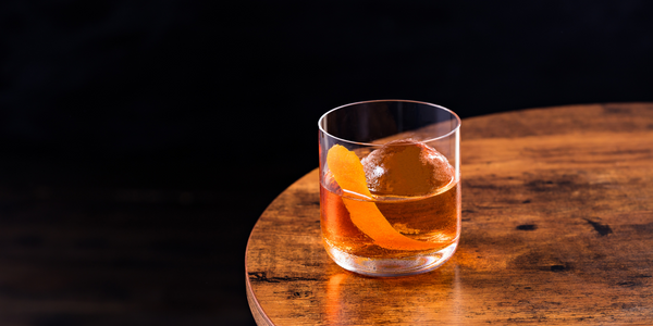 Photo of a whiskey old-fashioned over a sphere of ice, in a glass on a round wooden table against a black background