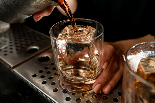 A rum old-fashioned is poured out of a metal mixing glass and into an old-fashioned glass with a large format ice cube held by a light brown-skinned hand with peach painted nails.
