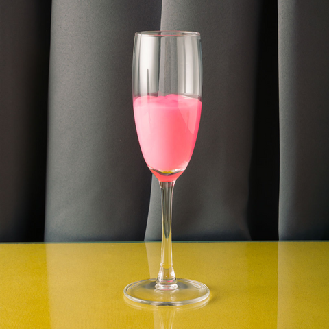 Champagne flute of something opaque pink against a grey velvet curtain background on a mustard yellow table 