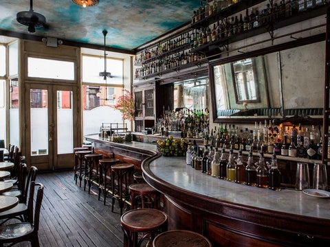 Photo of the bar at Hotel Delmano, Brooklyn, showing a wrap around white marble bar, dark wood stools and tables, an antique mirror above the backbar, and a ceiling painted with a blue sky and fluffy clouds