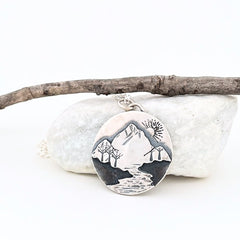 round silver pendant with mountains, a river and trees