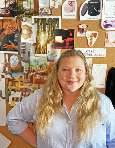 Chelsea Mooney, Founder of Terrae Collective, standing in front of her inspiration board full of images of nature, meditation, product references and home interiors.  She has long, wavy blond hair and is wearing a light blue oxford buttondown shirt.  She is smiling softly with one hand on her hip.