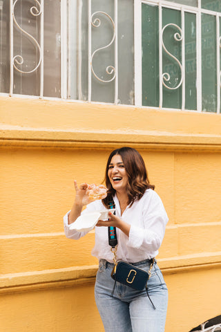 Jacqui Toumbas Nutritionist and Cook in white shirt and jeans, holding a pastry in front of an orange wall, laughing 