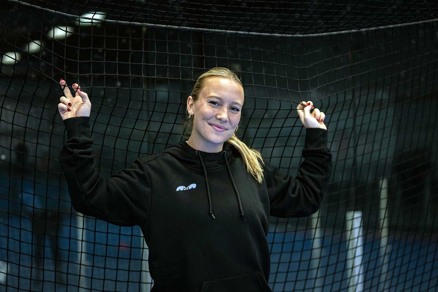 Field hockey player, Phia, wearing a TK sweatshirt with her hands up on a net.