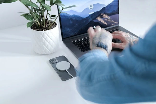 iPhone  avec un chargeur magsafe sans fil dessus et une personne en train d'utiliser un MacBook posé sur un bureau blanc avec une plante à côté