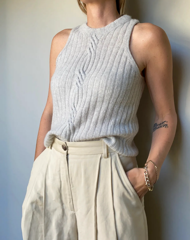 Shot of woman’s torso, wearing grey cabled knitted vest with hands in pockets of cream pleated trousers