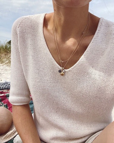 Close up of a woman wearing a white v-neck knitted tshirt with cluster of necklaces and beach scene behind