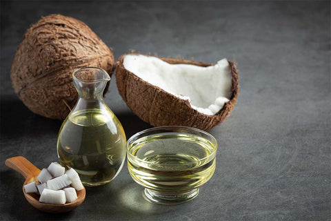 A still life of coconut oil products, including a whole coconut, halved coconut, oil in a glass jug and bowl, and chunks on a wooden spoon against a dark background.