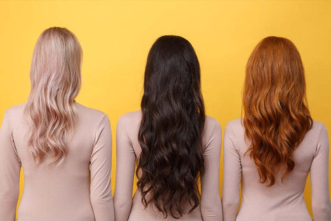 Three women facing away, displaying hairstyles in blonde, brunette, and auburn shades, each likely held with hair spray, against a vibrant yellow backdrop.
