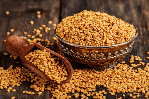 a bowl full of golden-brown fenugreek seeds, known for their benefits in hair oil formulations, placed on a rustic wooden background. A scoop is also filled with the seeds, and some are scattered around, highlighting the natural ingredient in its raw form, typically used for strengthening and nourishing hair.