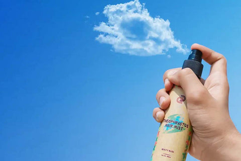 image shows a hand holding a bottle of "THEOPHRASTUS HAIR MIST" by Beauty Bliss against a clear blue sky with a fluffy cloud.