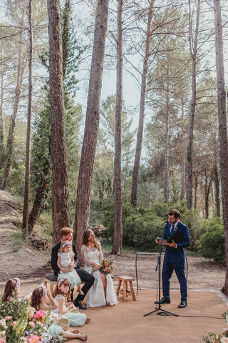 Photograph of the bride and groom sitting with girls, attentively listening to a man standing speaking to them through a microphone