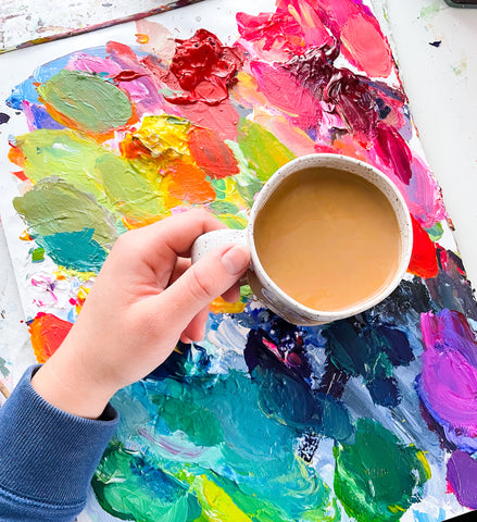 Artist's hand holding coffee cup on top of a painting palette.