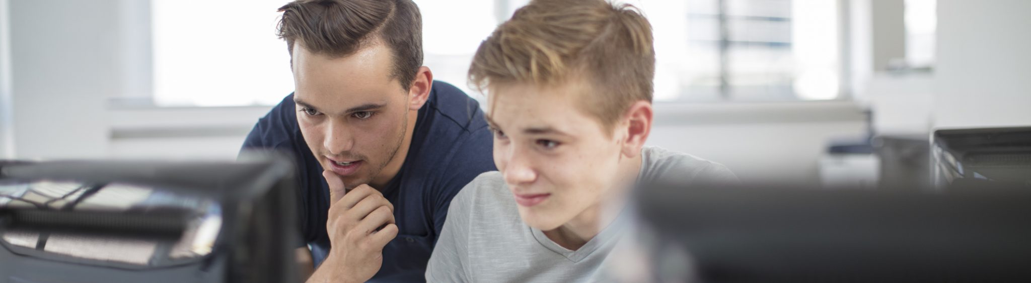 Teacher stands behind male student at desk with laptop open, ready to give instruction.