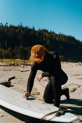 man wearing alki hat waxes a surf board