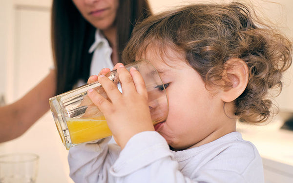 Ett barn dricker ett glas med appelsinjuice, mamman i bakgrunden