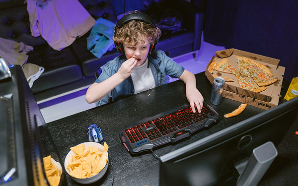 pojke i hörlurar spelar spel på datorn och äter kolhydrater i form av chips och pizza