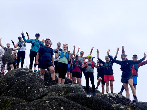 a large group of trail runners stand on top a mountain peak smiling at the camera which is at a lower angle than the runners.