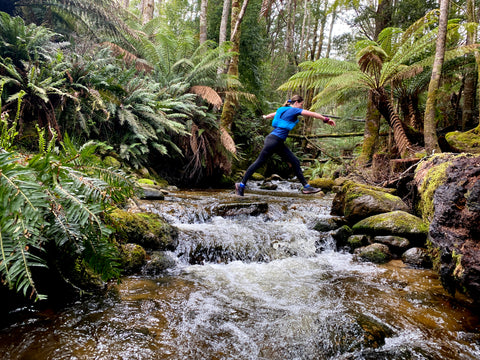 A woman wearing trail running attire and a bright blue vest pack is picked leaping over a creek, surrounded up ferns in the Tasmanian rain forest