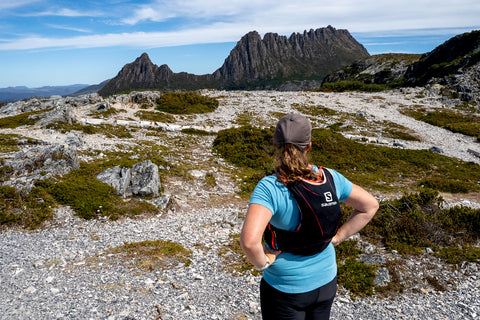 A female trail runner looks out across a chasm to view Cradle Mountain opposite her. It is a clear day with just a few white clouds behind the mountains peaks.