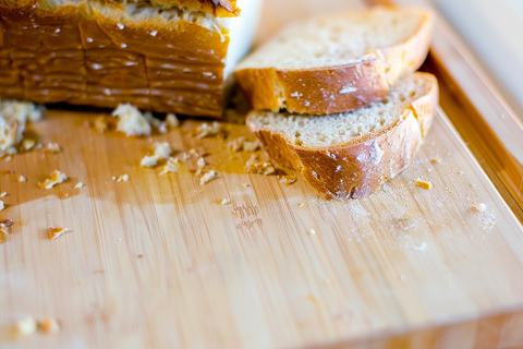 Close-up of a bamboo bread box cutting board with a partially sliced loaf of bread and collected crumbs in a built-in crumb catcher, showcasing its functionality and aesthetic appeal.