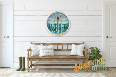 Sunshine Corner's Custom Lake House Sign That Says "Blue Skies and Dragonflies - Lake Life" hanging above a wood bench with pillows on it and greenery beside it.