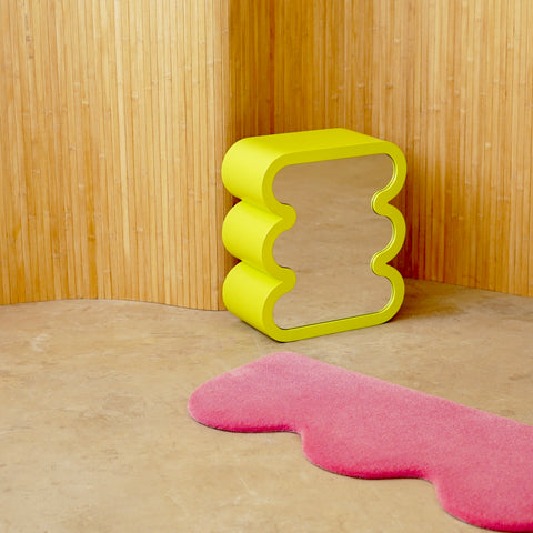 A stand alone mirror in color lime that is displayed on the ground together with a pink rug. 