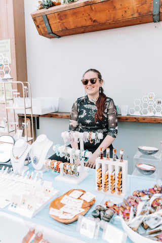 Lauren Lo Cornell, the artist and founder of Think Unique, sitting behind her display table filled with jewelry, essential oils, smudge bundles, crystals, and more. All displayed on a white airy backdrop with blue accents