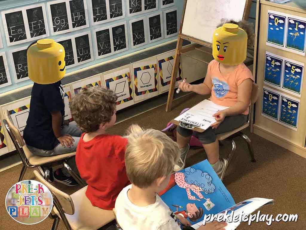 Students in pretend school dramatic play. One is a teacher is teaching preschool and taking attendance. The other kids are pretending to be her students.