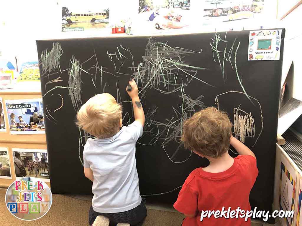 Kids are teaching preschool by writing on a pretend play chalkboard. 