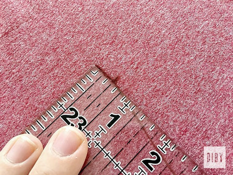 Fingers holding clear ruler on wrong side of pink knit fabric lining up markings.
