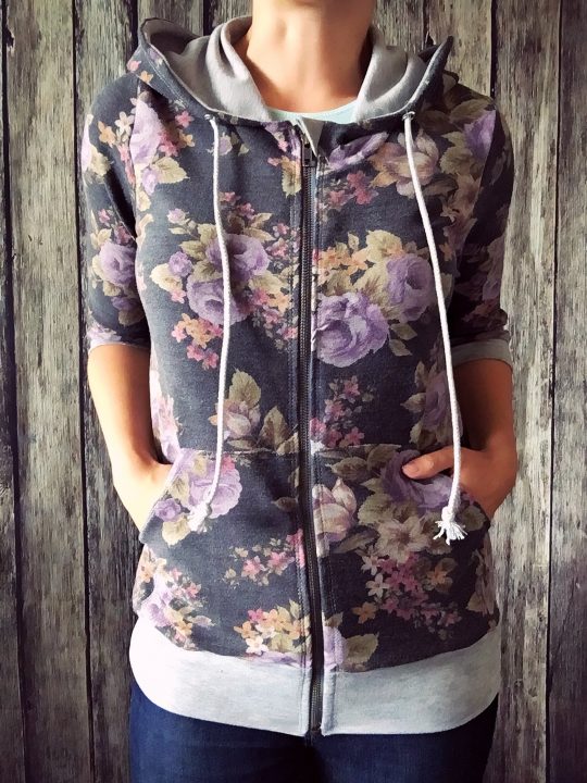 Picture of woman standing in front of a wall wearing a zipper front dark floral hoodie with gray trim and pockets.