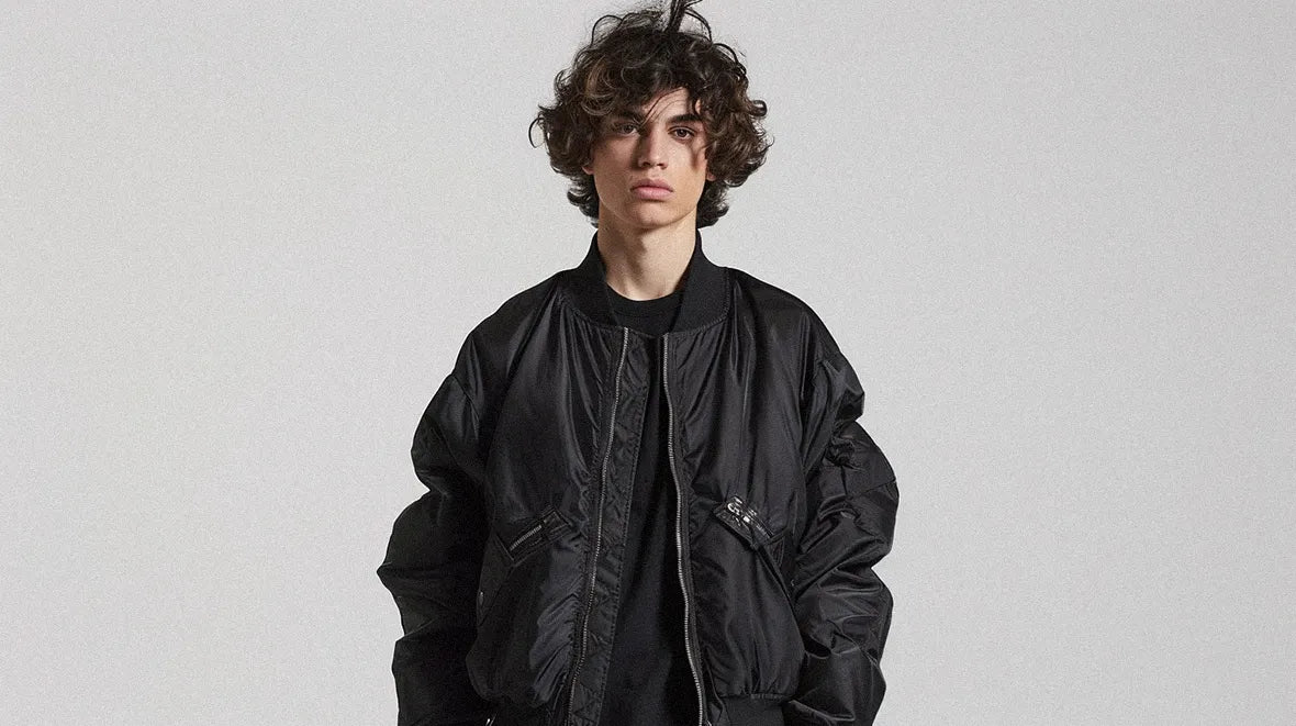 A person with curly hair models a black bomber jacket against a plain background.