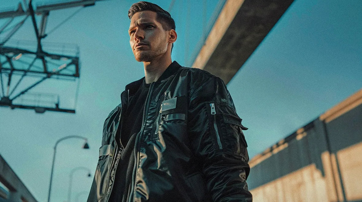 A person with a serious expression poses on a city street, dressed in a black techwear bomber jacket.