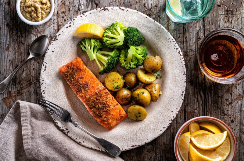 Cooked salmon, roasted potatoes, broccoli with mustard, drink, and lemon wedges on a rustic wooden table.