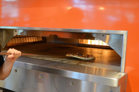 A person uses a long-handled peel to slide a pizza into a large, industrial oven with an orange exterior.