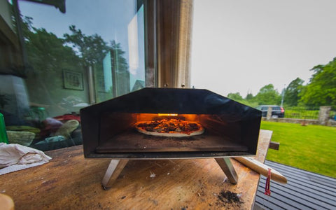 Portable outdoor pizza oven with a visible fire, set on a wooden deck with trees and a glass door in the background.