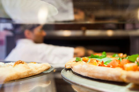 A chef in a white uniform placing a pizza into an oven, with freshly baked pizzas topped with tomatoes and basil in the foreground.