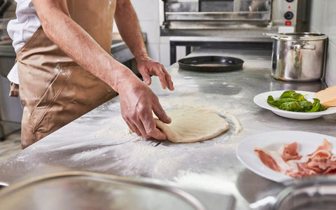 A person rolling out pizza dough on a floured kitchen counter.