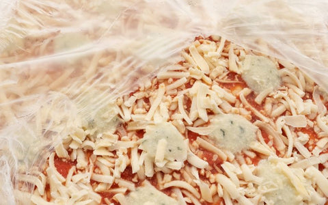 Close-up of a frozen pizza with shredded cheese and pieces of herb topping, partially obscured by plastic wrapping.
