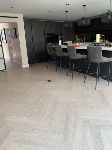 An image showcasing a sleek and modern room fitted with SPC (Stone Plastic Composite) flooring, warmed by underfloor heating. The room's large glass windows and bifold doors provide a contrasting view of a snowy, chilly outdoor scene, underscoring the inviting warmth of the indoor environment.