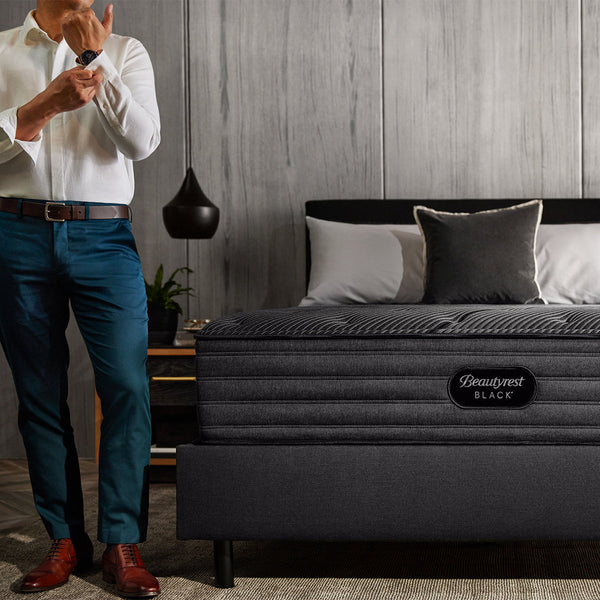 Man Buttoning Shirt Sleeve Standing Next To A Beautyrest Black L-Class Medium Mattress In Bedroom